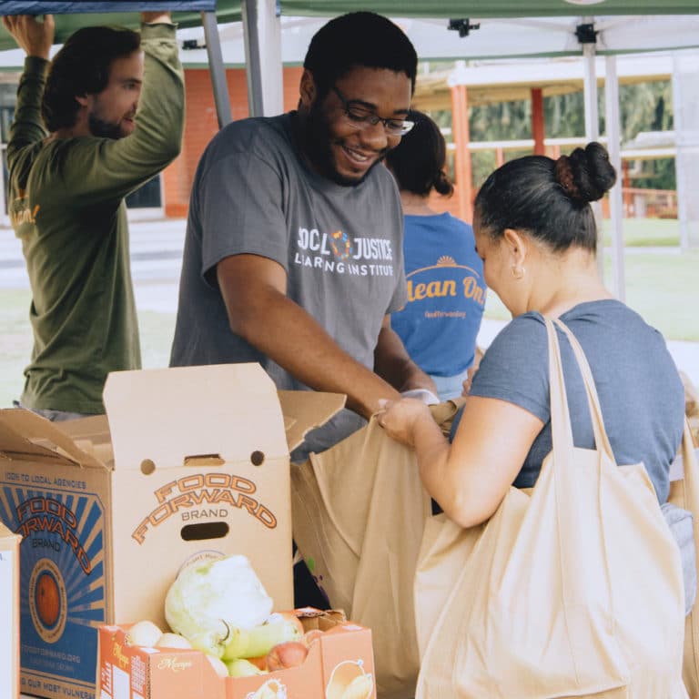 A volunteer hands a community member a bag of free produce at an outdoor Produce Pick-Up event.