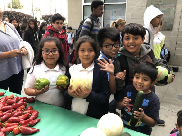 Five young children wave and hold up different fruits and vegetables at a Food Forward produce distribution.